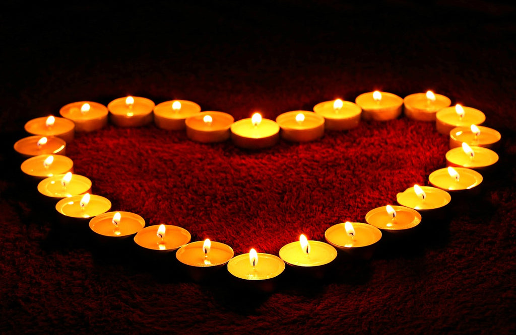 On a black background sits a bunch of tea light candles in the shape of a heart all lit and burning brightly in colours of orange and yellow.