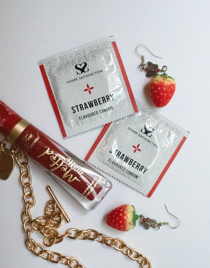 Two foil square packets lay on a white table surrounded by a pair of strawberry earings, a gold chain and a red lip balm.