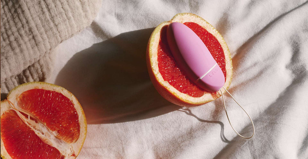 An orange cut in half laying face up on a bed and one slice has a pale pink bead laying on top of it that has a string tail.