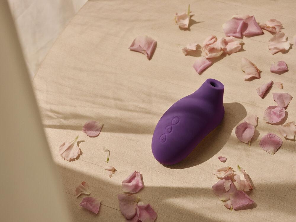 The corner of a bed covered in a pale coloured linen sheet sprinkled with pink rose petals with a purple rubber item in the middle with a bloated belly and a small suction head. 