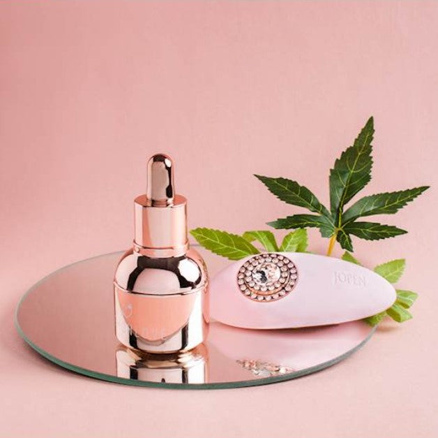 On a pale pink background sits a mirrored tray and behind this a a couple of green leaves and a shiny bottle in the foreground with a dropper lid and a pale pink rubber looking oval object behind the bottle.