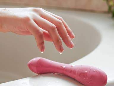 A woman's hand reaching from out of the bath tub to the edge of the tub with dripping wet fingers reaching for a long pink rubber looking object.