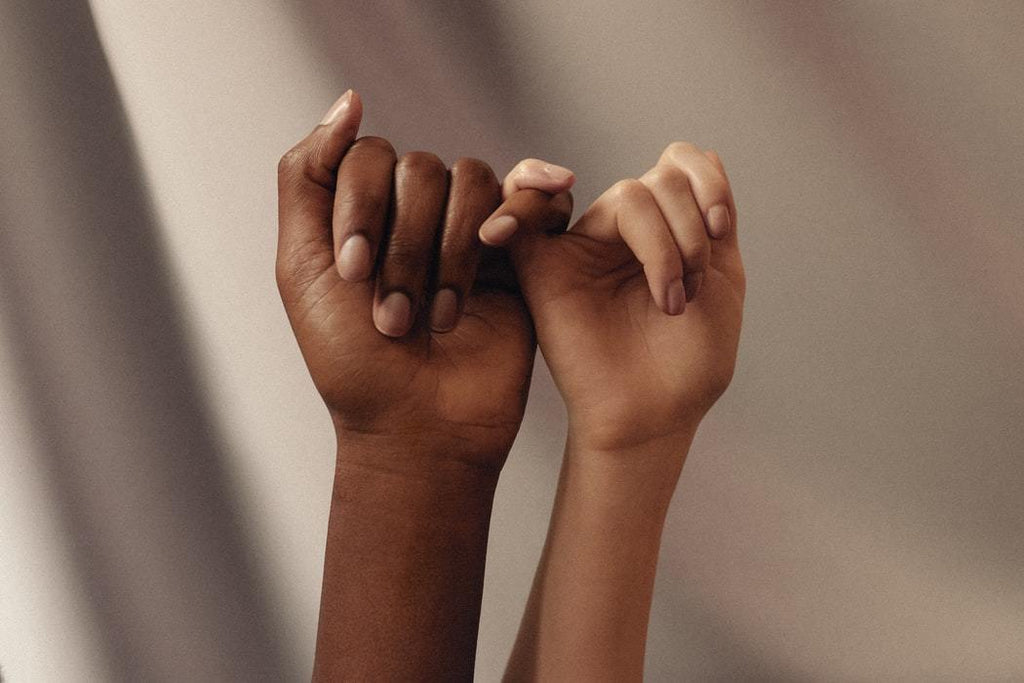 Two woman's well manicured hands holding on to each other using their pinky fingers only and in two different skin tones.