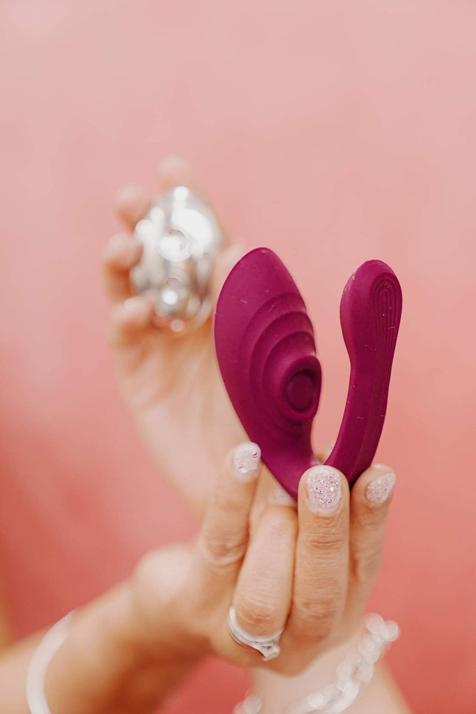 A woman's hand is in the foreground with glitter nail polish and holding a dark pink rubber V shaped item with an 'eye' on one side. The woman's other hand is in the background, out of focus, holding something shiny in front of a pale pink background.