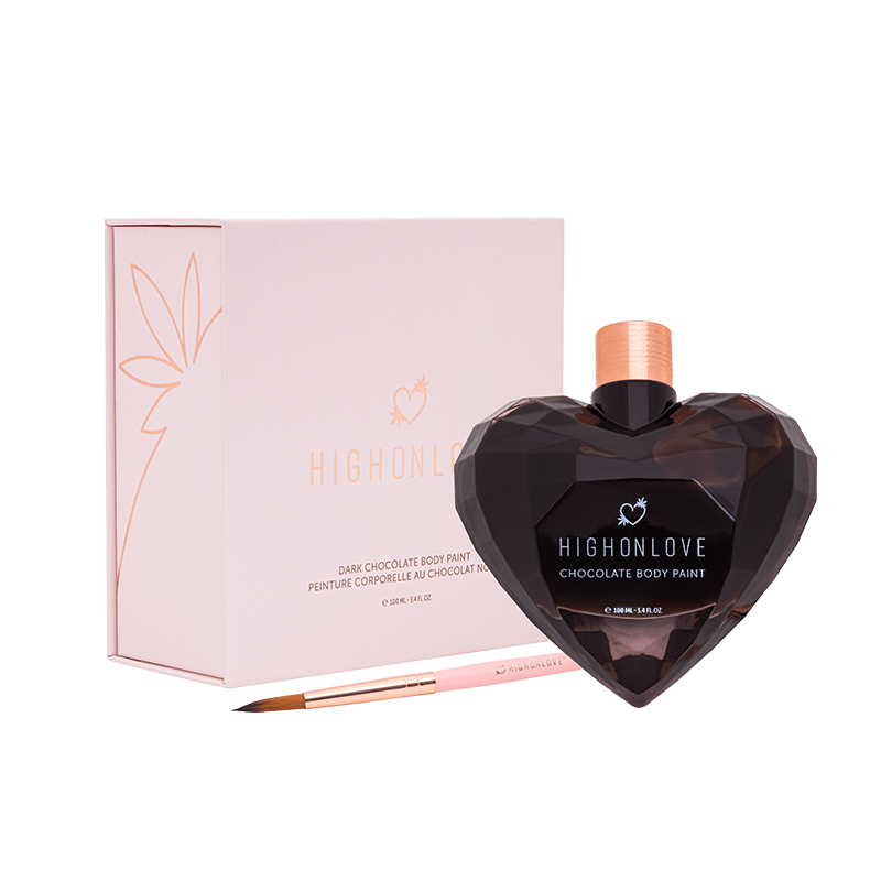 Brown heart shaped bottled resembling a perfume bottle with a shiny gold lid sitting beside a delicate looking paint brush and pale pink box.