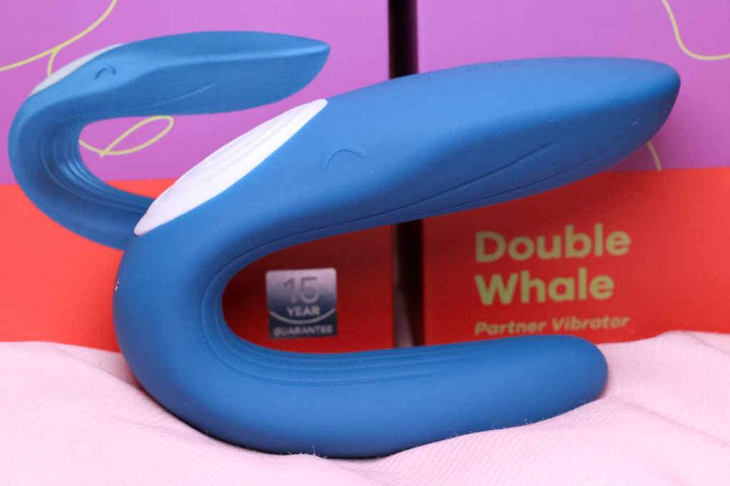 Blue rounded product with a white hat and nubbed eyes resembling a smiling whale sitting on a pale pink silk sheet with the box displayed behind.