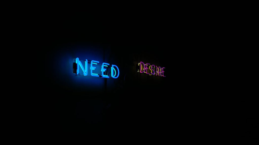 A black image with two neon signs pictured in the middle. One is the word need lit up in blue and the other is the word desire lit up in red.