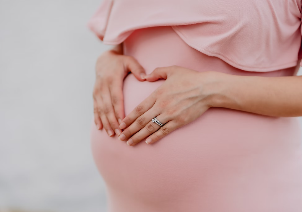 A woman's torso pictured up close and she is wearing a tight fitted pale pink top and is holding both hands on her pregnant stomach with her fingers making a heart shape.