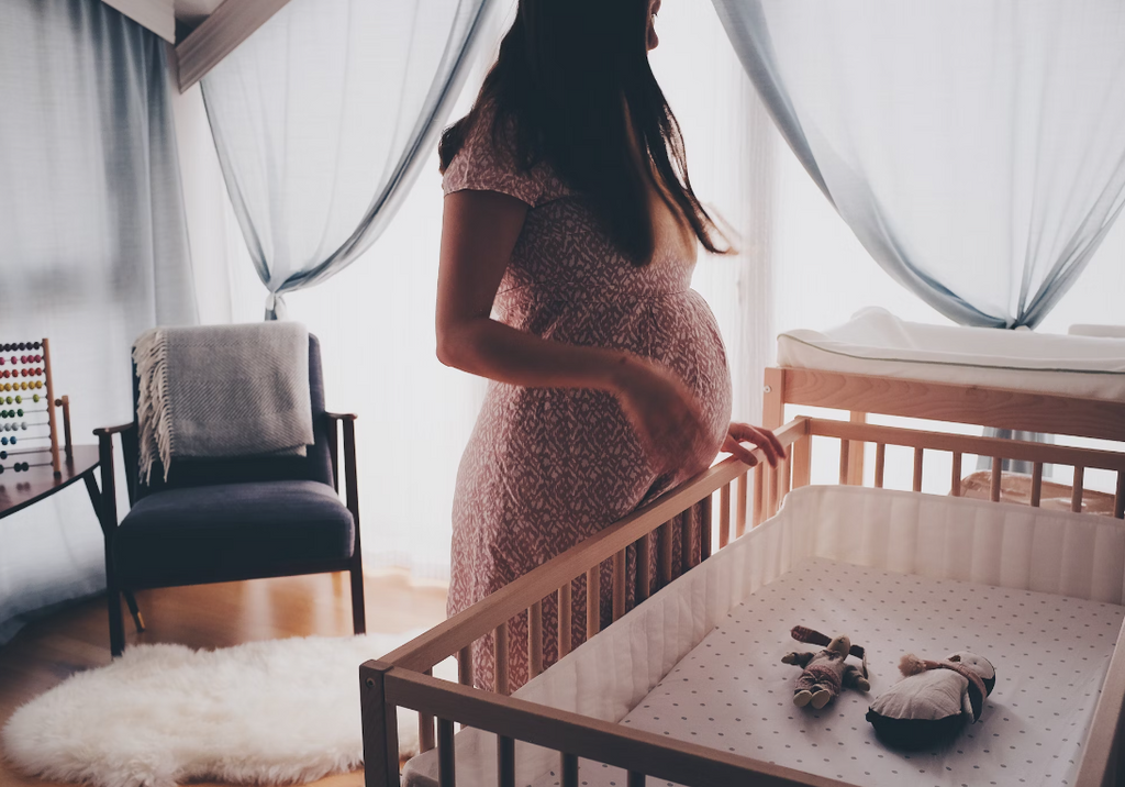 A woman is pictured in a small bedroom overlooking a baby's cot. She is wearing a long dress and has a pregnant stomach. The bedroom windows are covered with white sheer curtains and there is a small chair placed in one corner of the room.