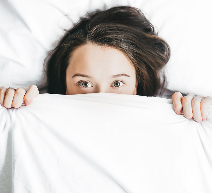 A woman is pictured at close range laying in bed on white sheets with her brown hair out. Only half her face is visible as she has the white bedspread pulled up to her nose and her eyes are staring straight ahead.