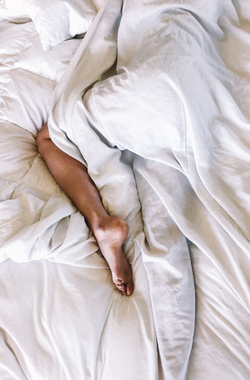 A zoomed in photo of a woman laying on a mattress with white sheets and she is visible from the waist to her toes and has one foot outside of the white sheet while the rest of her body is covered.